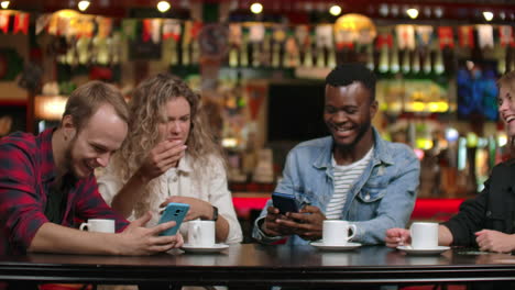 Four-friends-sitting-in-a-cafe-laugh-and-smile-while-looking-at-the-screens-of-phones-and-exchanging-photographs-from-travel.-African-American-girlfriend-chatting-and-drinking-coffee.-Multinational-company-of-friends.